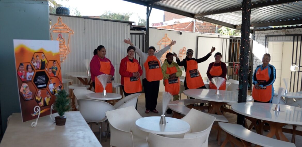 Seven people wearing orange aprons stand and wave inside a covered outdoor seating area with white tables and chairs. A small potted plant and a colorful poster are visible in the foreground.