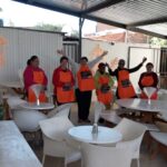 Seven people wearing orange aprons stand and wave inside a covered outdoor seating area with white tables and chairs. A small potted plant and a colorful poster are visible in the foreground.
