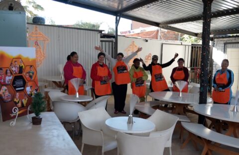Seven people wearing orange aprons stand and wave inside a covered outdoor seating area with white tables and chairs. A small potted plant and a colorful poster are visible in the foreground.