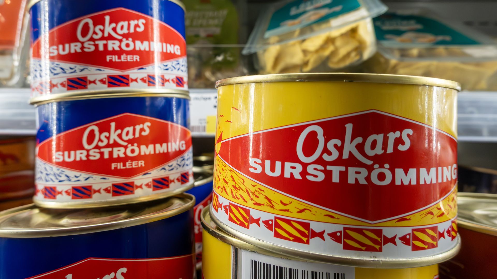 Close-up of stacked cans of Oskars Surströmming, a Swedish fermented fish with a tang reminiscent of vinegar, featuring colorful red, yellow, and blue labels on a store shelf. Other packaged food is blurred in the background.
