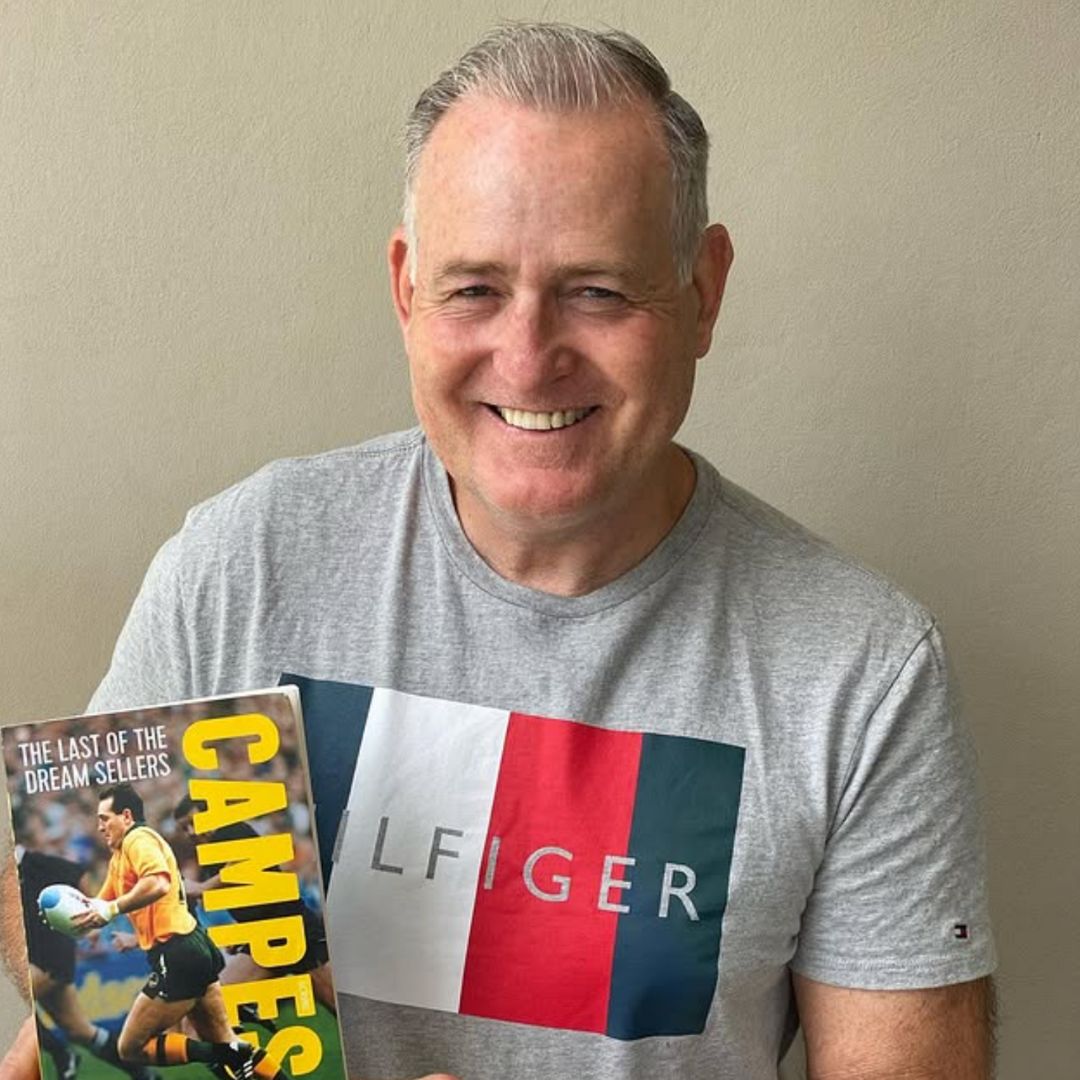 A smiling man with short gray hair wears a gray Tommy Hilfiger T-shirt and holds up a rugby book titled CAMPESI: The Last of the Dream Sellers with a photo of a rugby player on the cover.