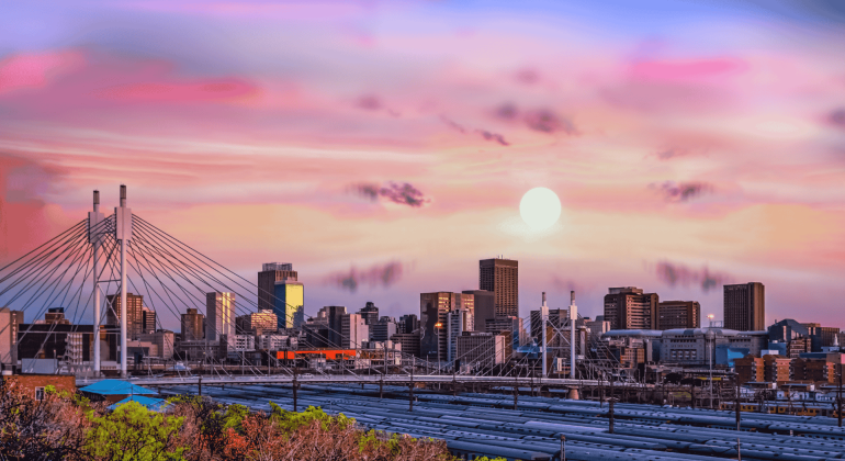 A vibrant Joburg city skyline at sunset, featuring a modern cable-stayed bridge in the foreground and tall buildings under a colorful sky with pink, purple, and orange hues. The sun is low on the horizon.