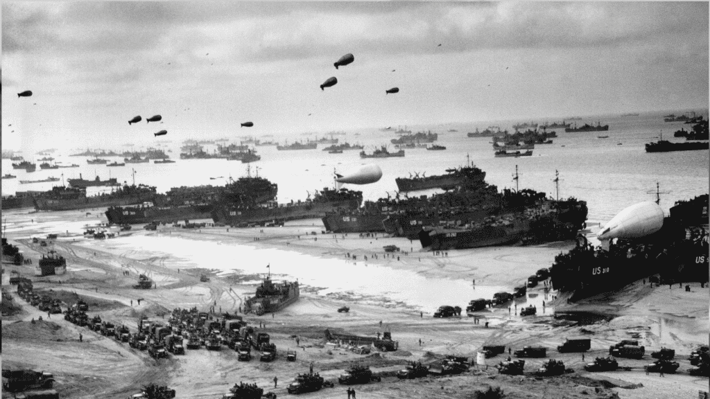 A black-and-white photo shows numerous military ships and landing craft at a crowded beach during World War II. Vehicles, troops, and equipment fill the shore, while Spitfire fighters soar above barrage balloons and many ships offshore.