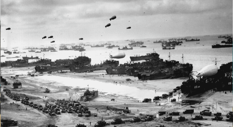 A black-and-white photo shows numerous military ships and landing craft at a crowded beach during World War II. Vehicles, troops, and equipment fill the shore, while Spitfire fighters soar above barrage balloons and many ships offshore.