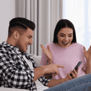 A man in a checkered shirt and a woman in a pink shirt, embodying Gen Z energy, sit on a couch looking at a phone. He points at the screen while she smiles excitedly, hands raised in surprise—capturing the spirit of the hustle.