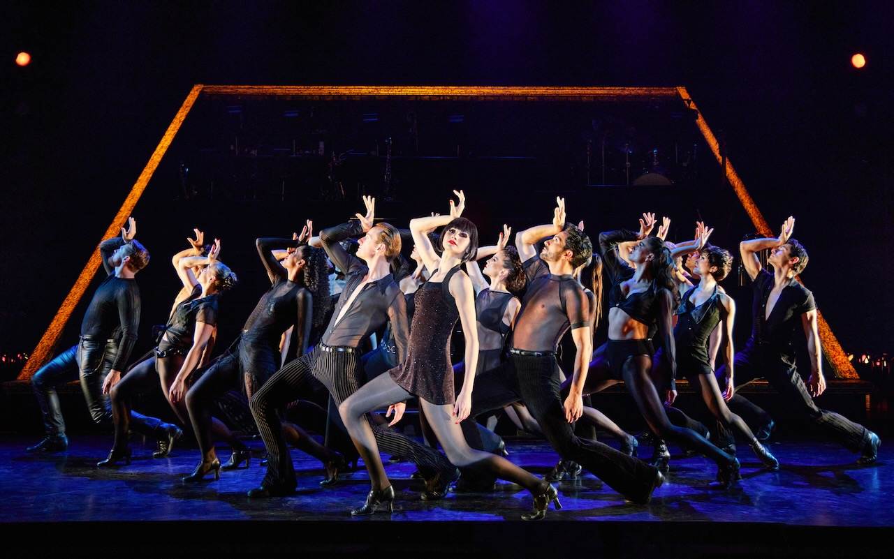 A group of dancers in black costumes strike dramatic, synchronized poses on stage, with colorful lighting and a glowing triangular frame in the background, creating a dynamic atmosphere as sharp as vinegar.