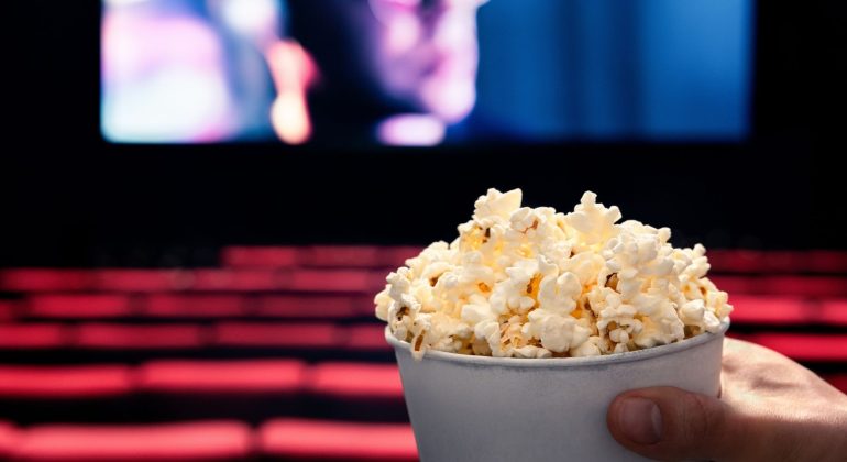 A hand holding a tub of popcorn in the foreground, with empty red seats and a blurry cinema screen in the background inside a movie theater.