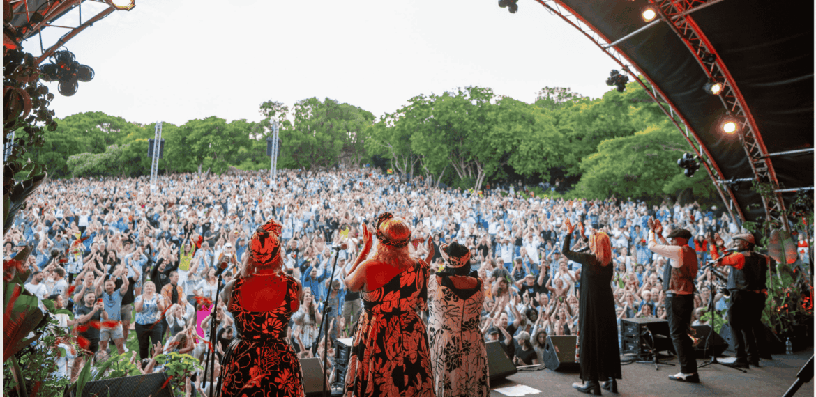 A group of performers on an outdoor stage face a large, enthusiastic crowd with raised hands, surrounded by lush green trees under a partly cloudy sky.