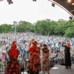 A group of performers on an outdoor stage face a large, enthusiastic crowd with raised hands, surrounded by lush green trees under a partly cloudy sky.