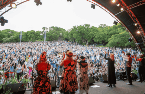 A group of performers on an outdoor stage face a large, enthusiastic crowd with raised hands, surrounded by lush green trees under a partly cloudy sky.