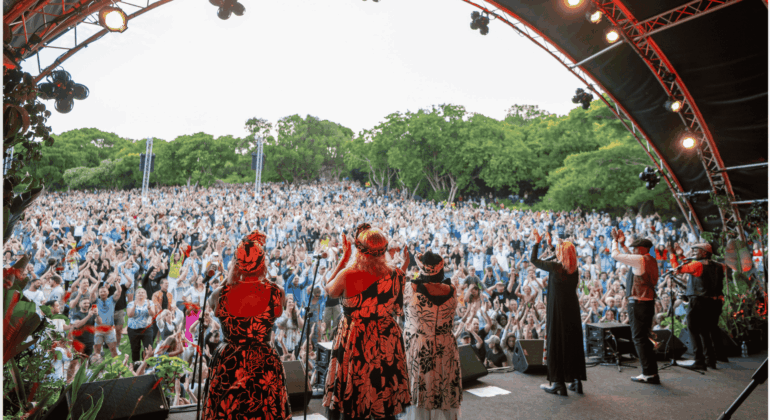 A group of performers on an outdoor stage face a large, enthusiastic crowd with raised hands, surrounded by lush green trees under a partly cloudy sky.