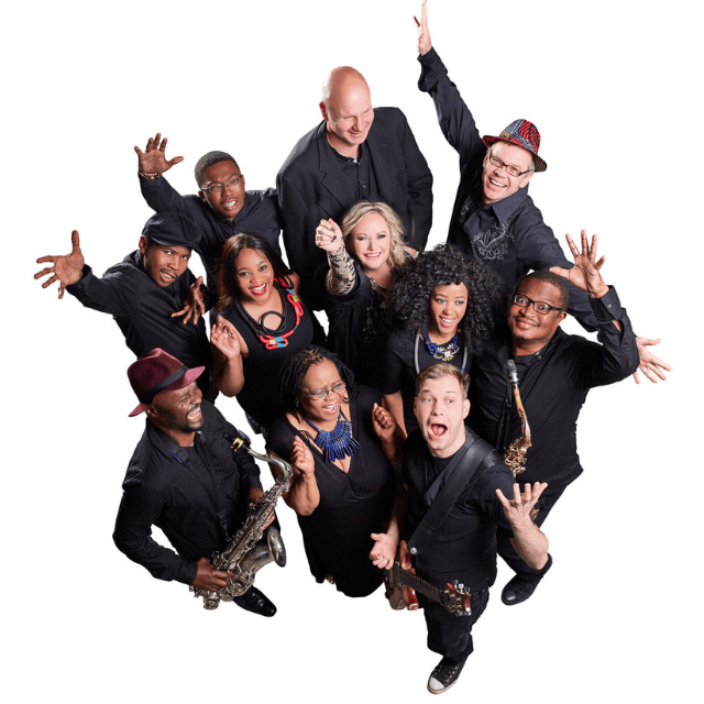 A diverse group of eleven musicians dressed in black, holding instruments and smiling enthusiastically, pose together against a white background, appearing joyful and energetic.