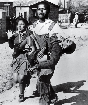 A young man runs while carrying an injured child in his arms, with a distressed girl beside him. The scene unfolds on a street lined with houses, other people, and scattered mixtapes in the background.
