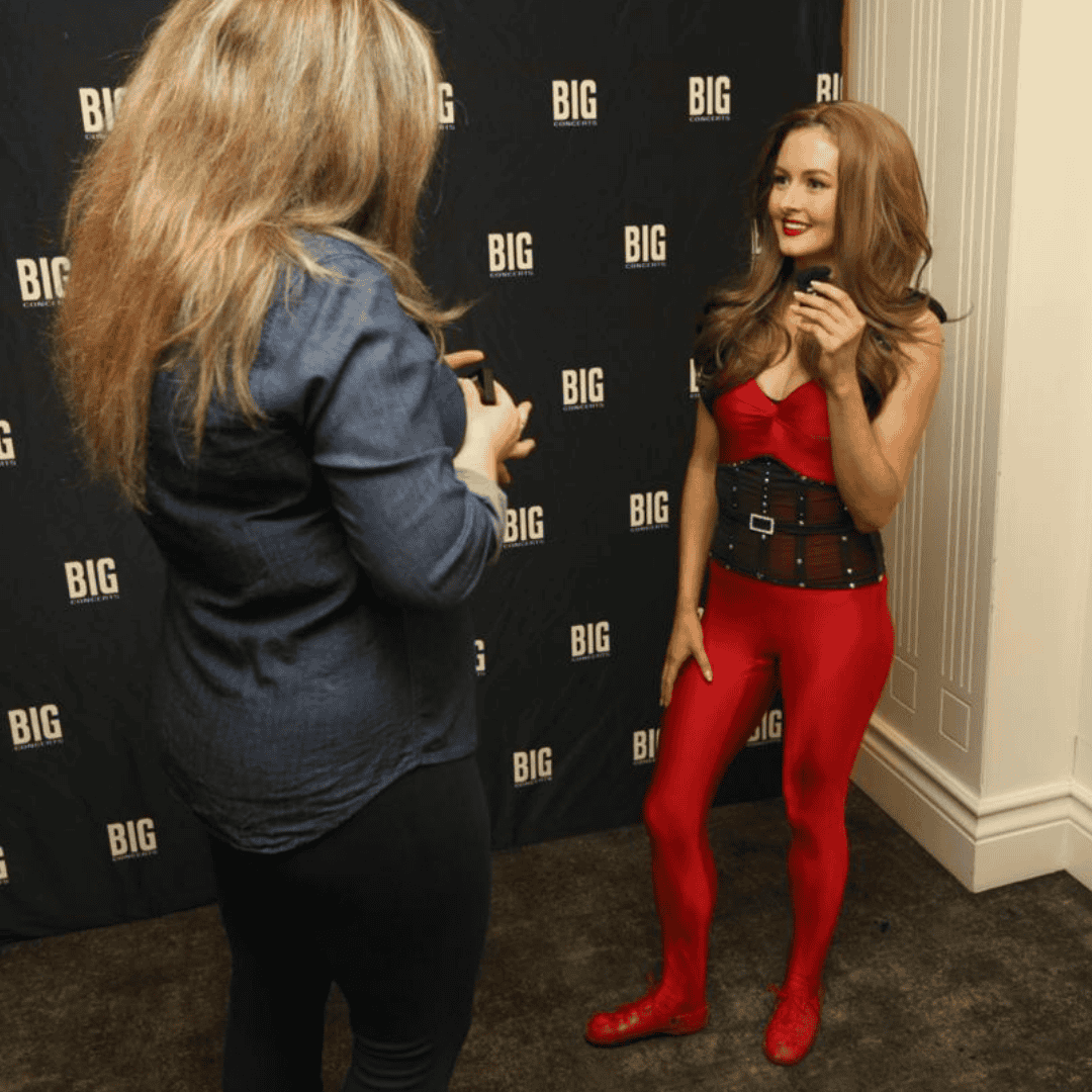 A woman in a red costume with boots and a wide belt poses and smiles, channeling Lord of the Dance energy, while another person with long hair and casual clothes prepares to take her photo in front of a BIG step-and-repeat backdrop.