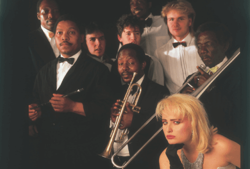 A group of eight men in tuxedos, some holding brass instruments, pose together against a dark background. In the foreground, a blonde woman in a white dress and black gloves sits, looking intently at the camera.