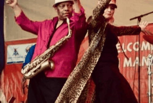 A man in a white hat playing a saxophone stands beside a woman in a long cheetah-print coat, both with raised arms, performing energetically on an outdoor stage with a red backdrop.