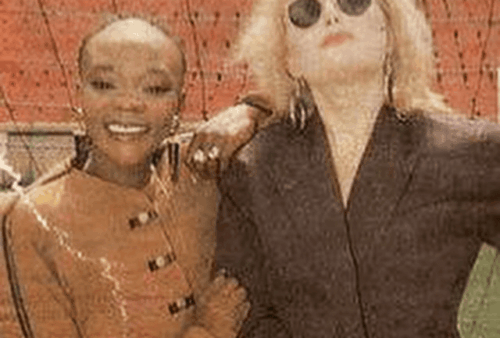 Two women pose together outdoors in front of a brick wall. One woman wears a tan jacket and smiles, while the other wears dark sunglasses and a dark outfit, looking up with a relaxed expression.
