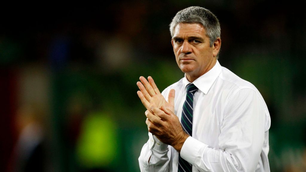 A man, resembling Nick Mallet, in a white dress shirt and striped tie stands looking serious with his hands clasped together. The background is blurred with green and dark tones.