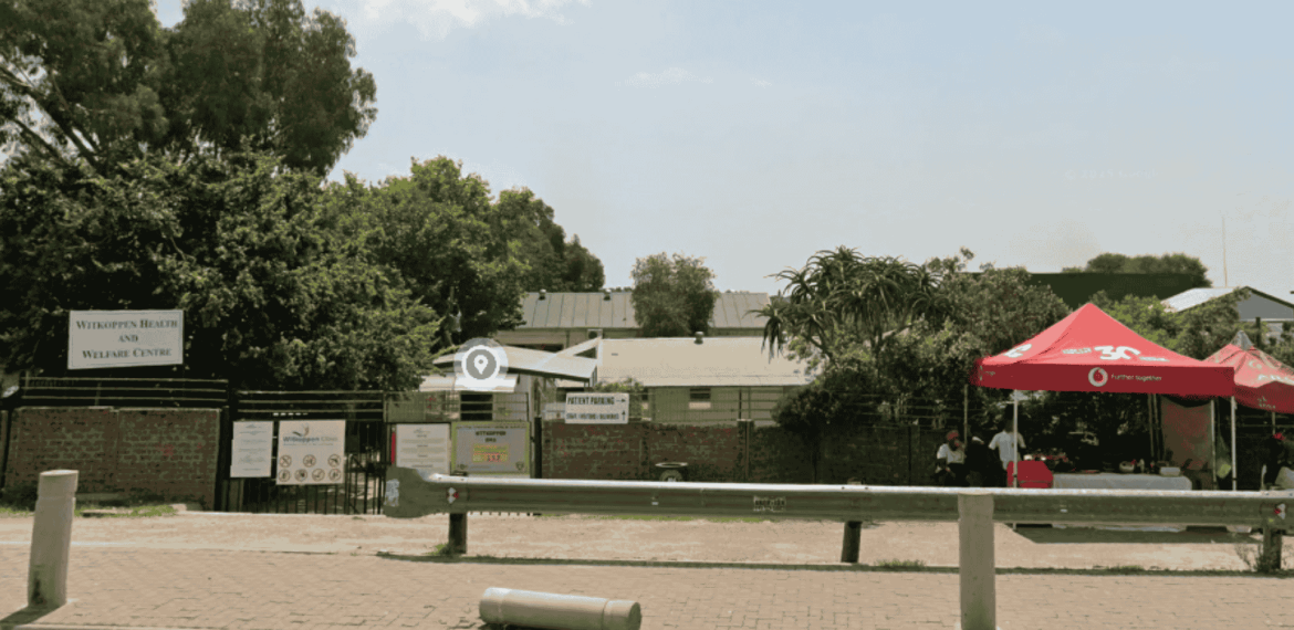 A paved parking area in front of a fenced property near Witkoppen Clinic features signs, a red tent with two people seated beneath, and is bordered by trees, greenery, and a metal barrier along the edge.