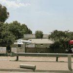 A paved parking area in front of a fenced property near Witkoppen Clinic features signs, a red tent with two people seated beneath, and is bordered by trees, greenery, and a metal barrier along the edge.