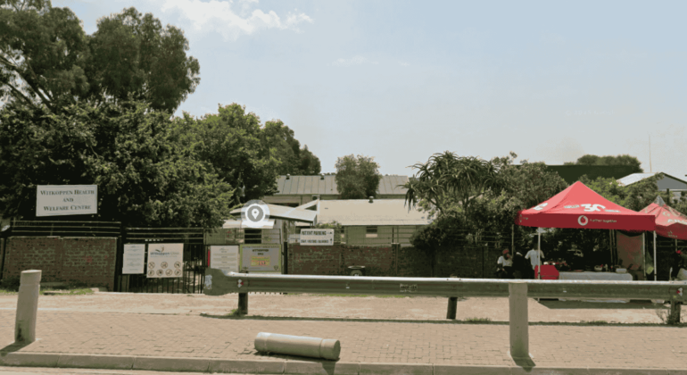 A paved parking area in front of a fenced property near Witkoppen Clinic features signs, a red tent with two people seated beneath, and is bordered by trees, greenery, and a metal barrier along the edge.