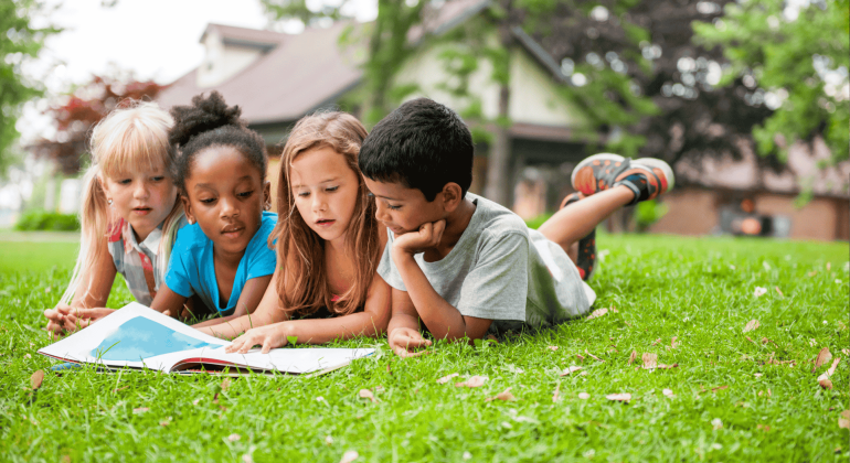 Four young children lie on the grass outside, closely reading a large open book together, imagining stories about the ict sector, with trees and a house in the background on a sunny day.