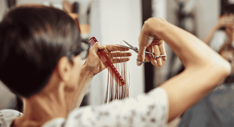 A hairstylist with short dark hair holds a red comb and scissors, carefully trimming long strands of hair in a salon, showcasing expert Hair Care. The focus is on the stylist’s hands and tools.