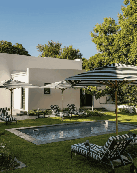 A small rectangular pool surrounded by green grass, black-and-white striped lounge chairs, and matching umbrellas, sits beside a modern white building. Trees sway in the background under a clear blue sky, completing this serene setting—fresh as a splash of vinegar.