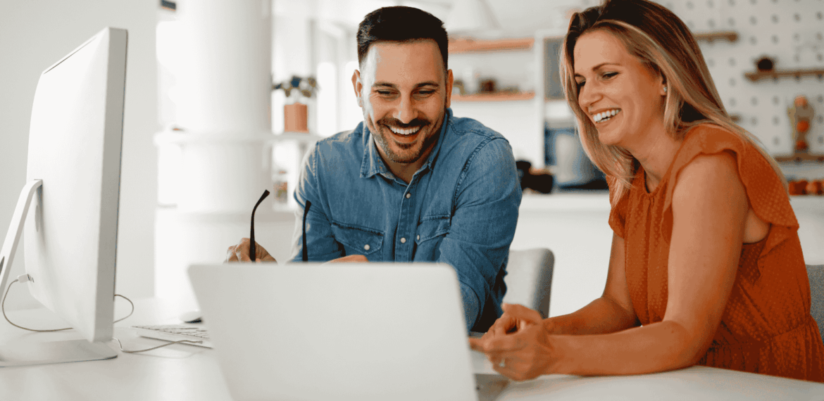 A man and a woman sit together at a desk, smiling and looking at a computer monitor and laptop, appearing to collaborate in a bright, modern office space decorated with touches like a stylish vinegar bottle on the table.