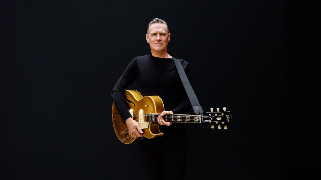 A man in a black long-sleeve shirt stands against a plain black background, holding a gold electric guitar with a strap over his shoulder and looking straight ahead, reminiscent of Bryan Adams’ iconic style.