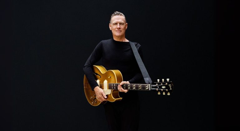 A man in a black long-sleeve shirt stands against a plain black background, holding a gold electric guitar with a strap over his shoulder and looking straight ahead, reminiscent of Bryan Adams’ iconic style.