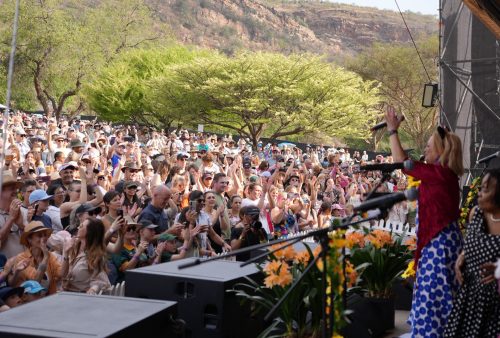 A performer on an outdoor stage entertains a large, cheering crowd. Fans in hats and sunglasses enjoy the sunny day, as Dave Matthews music fills the air amid trees, mountains, and vibrant festival decorations in the background.