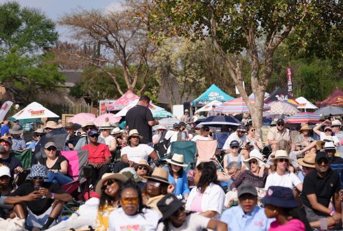 A large, diverse crowd sits outdoors on chairs and blankets at a festival or Dave Matthews event, shaded by hats and umbrellas. Colorful tents and trees are visible in the background under a sunny sky.