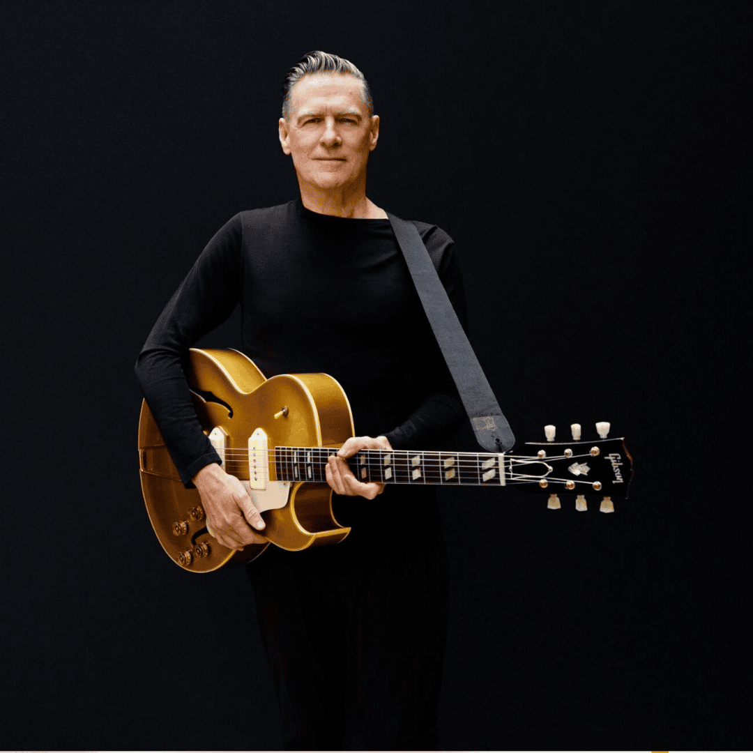 A man with short gray hair, wearing a black long-sleeve shirt, stands against a plain dark background holding a gold electric guitar with a black strap across his shoulder, resembling Bryan Adams.