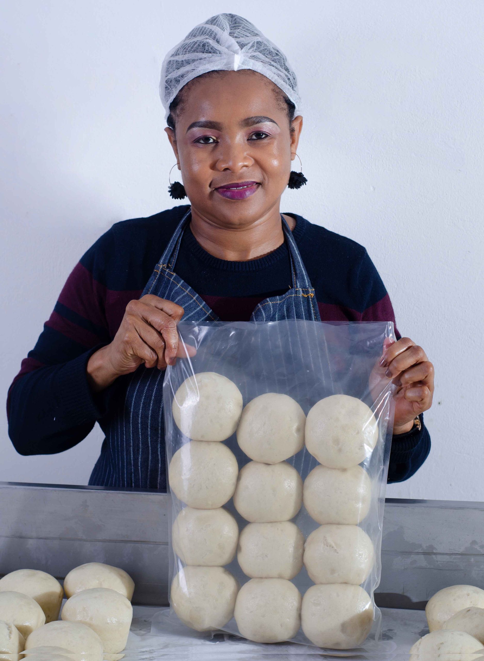 A smiling foodie in a hair net, dark sweater, and apron holds a clear bag with twelve round dough balls. More dough balls are on the table in front of her as she stands against a plain white background.