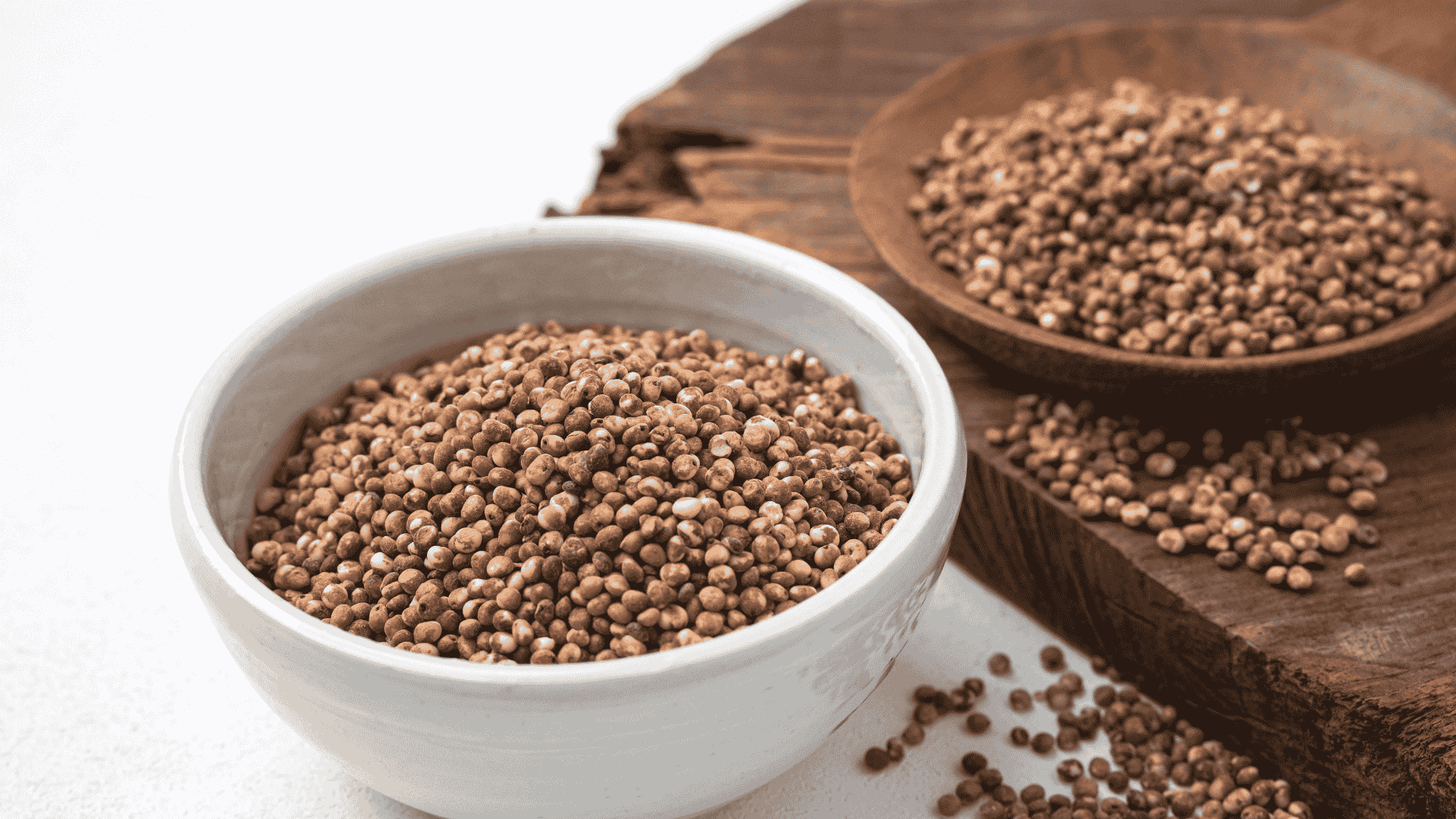 A white bowl filled with brown quinoa grains sits on a light surface next to a wooden plate holding more quinoa, with some grains scattered around—a simple yet inviting scene for any foodie.
