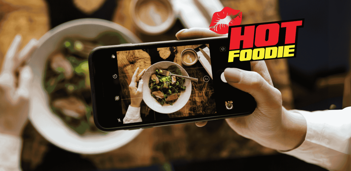 A foodie snaps a photo of a salad bowl with their smartphone. The words “HOT FOODIE” and a red lipstick kiss graphic appear on the right side. The table is set with delicious food and drinks.