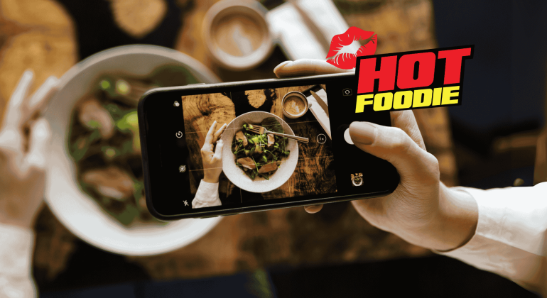 A foodie snaps a photo of a salad bowl with their smartphone. The words “HOT FOODIE” and a red lipstick kiss graphic appear on the right side. The table is set with delicious food and drinks.