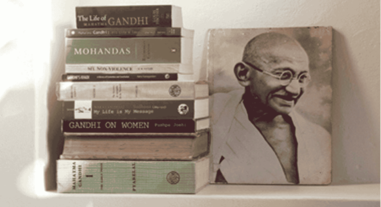 A stack of books about Mahatma Gandhi is arranged beside a black-and-white photograph of Gandhi smiling and wearing glasses and a traditional shawl, reminiscent of historic photos displayed at Witkoppen Clinic.