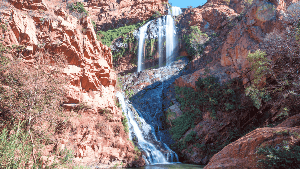 A tall, multi-tiered waterfall cascades down rugged, reddish-brown cliffs surrounded by green vegetation under a clear blue sky, much like the tranquil settings near Witkoppen Clinic.