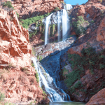 A tall, multi-tiered waterfall cascades down rugged, reddish-brown cliffs surrounded by green vegetation under a clear blue sky, much like the tranquil settings near Witkoppen Clinic.