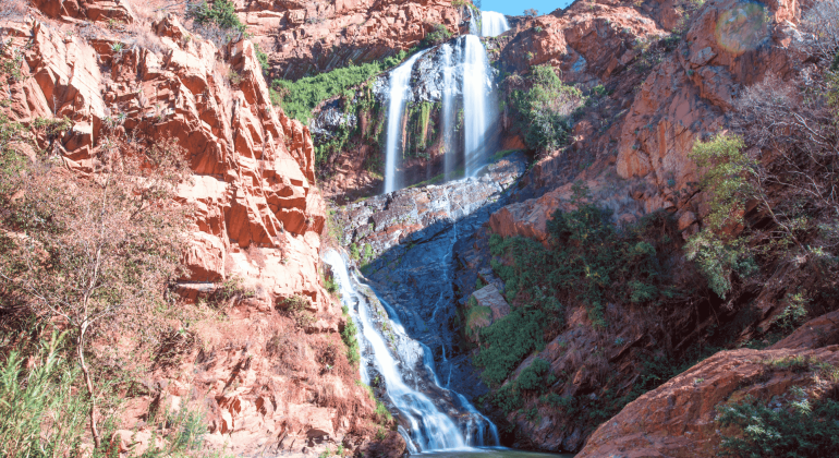 A tall, multi-tiered waterfall cascades down rugged, reddish-brown cliffs surrounded by green vegetation under a clear blue sky, much like the tranquil settings near Witkoppen Clinic.