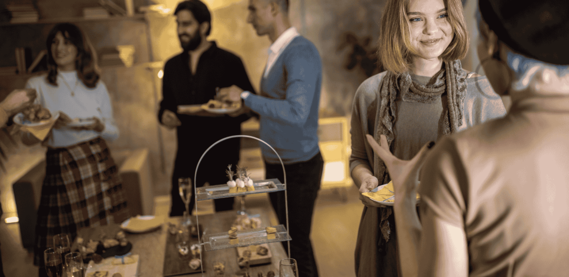 A small group of people at a cozy indoor party, chatting and smiling while holding plates of food, celebrate homeownership. A table with assorted appetizers and drinks is in the foreground.
