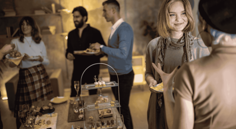 A small group of people at a cozy indoor party, chatting and smiling while holding plates of food, celebrate homeownership. A table with assorted appetizers and drinks is in the foreground.