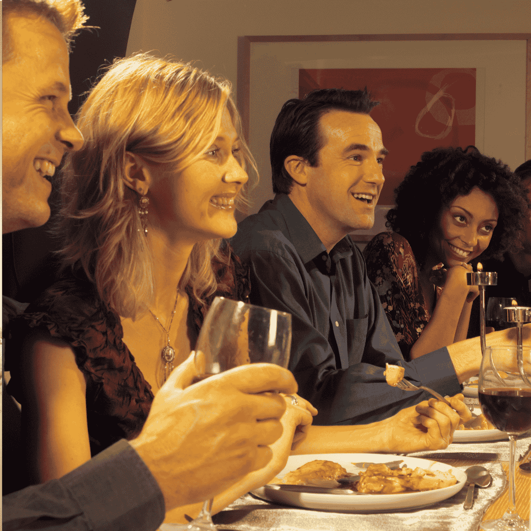 Four people sit closely together at a dinner table, smiling and laughing while enjoying food and drinks. The warm, friendly mood hints at the joy of homeownership, with plates, glasses, and utensils visible in front of them.