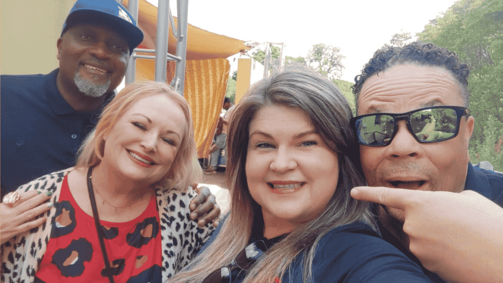 Four adults smiling and posing together outdoors. The woman in front is taking the selfie, while a man on the right points toward her. Bright sunlight and greenery set the scene for this Dave Matthews-inspired gathering.