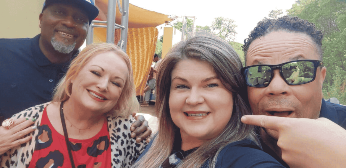 Four adults smiling and posing together outdoors. The woman in front is taking the selfie, while a man on the right points toward her. Bright sunlight and greenery set the scene for this Dave Matthews-inspired gathering.