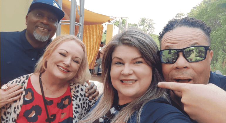 Four adults smiling and posing together outdoors. The woman in front is taking the selfie, while a man on the right points toward her. Bright sunlight and greenery set the scene for this Dave Matthews-inspired gathering.