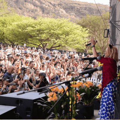 A performer in a blue and white dress sings on an outdoor stage before a large, enthusiastic crowd, reminiscent of a Dave Matthews concert. Lush green trees and mountains are visible in the background under a bright sky.