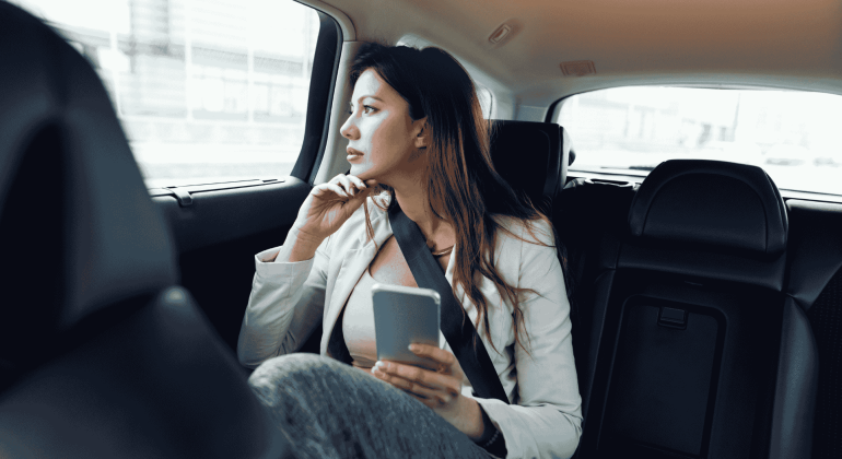 A woman sitting in the backseat of a car, wearing a seatbelt, holds a smartphone and gazes thoughtfully out the window, pondering her journey toward homeownership during the daytime.